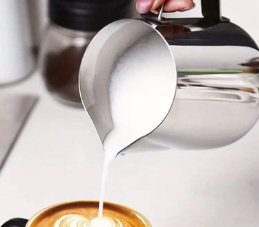 Stainless steel pitcher pouring milk into a coffee cup with a blurred background